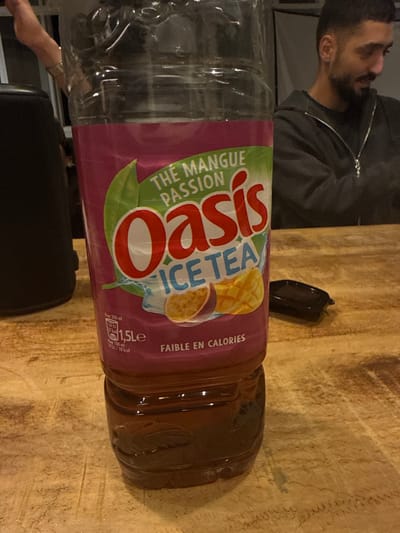 The image captures a dimly lit indoor scene in Saint-Denis, France, centered on a partially consumed 1.5-liter plastic bottle of Oasis Ice Tea, "Thé Mangue Passion," resting on a light-colored wooden table. The bottle's label, featuring mango and passion fruit illustrations, indicates "FAIBLE EN CALORIES" (low in calories) and contains a dark brown liquid, approximately one-quarter full. To the immediate right of the bottle on the table is a small, dark, rectangular container with a lid. In the background, a male individual with dark hair and a beard is visible, wearing a dark hooded sweatshirt with a zipper; he is seated, looking downwards and to his right, away from the camera. A partial view of a person's forearm and hand, adorned with a metallic bracelet, is visible in the upper left background, next to a dark, cylindrical object on the table. No direct interaction with the bottle is observed.