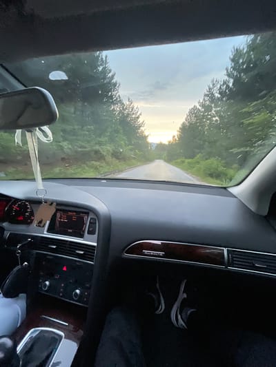 An interior shot from the perspective of a driver or passenger in a car, looking forward through the windshield. The scene is a paved road curving through a dense forest of tall evergreen trees. The sky above is a mix of pale blue and grey clouds, with the sun setting or rising in the distance, casting a soft glow on the horizon. The car's dashboard is visible, featuring an infotainment screen displaying "Radio FM" and other text, a tachometer, and various control buttons. A wooden charm hangs from the rearview mirror. The overall impression is a journey through a natural, wooded landscape at twilight. The image does not contain any discernible landmarks or features that would allow for identification of the specific location in Delchevo, Bulgaria.