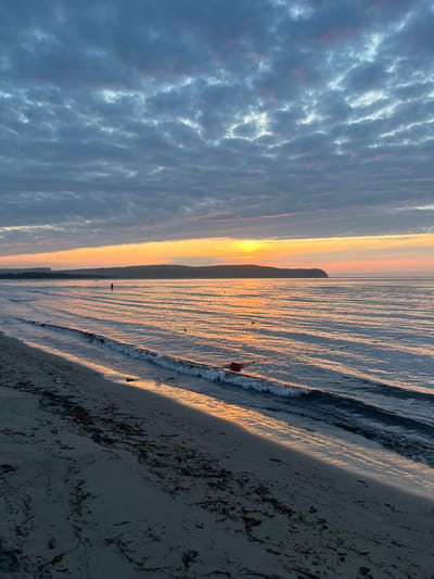 The image depicts a serene beach scene at sunrise in Datta, Russia. The sky is filled with dramatic, layered clouds, predominantly in shades of gray, with hints of pink and orange from the rising sun. The sun itself is a bright orb peeking through a break in the clouds, casting a warm, golden glow across the water and the distant hills. Gentle waves are rolling onto the sandy shore, creating ripples and reflections on the wet sand. Scattered along the shoreline and in the shallow water are small, dark objects, possibly seaweed or debris. In the distance, a lone figure stands in the water, silhouetted against the bright horizon. A small, red object, likely a buoy or marker, floats in the water closer to the shore. The beach itself is composed of dark sand with patches of seaweed and other organic material scattered across it. The overall impression is one of tranquility and the peaceful beginning of a new day.