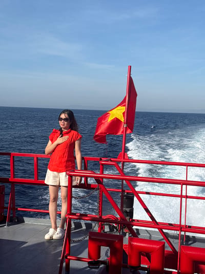 A woman stands on the deck of a boat, her left hand placed over her chest. She is wearing a red short-sleeved shirt, beige shorts, white clogs, and sunglasses. The boat has red railings and a red flag with a yellow star is flying from a pole on the right side of the image. The boat is in the ocean, with a wake trailing behind it. The sky is blue with a few faint clouds. The location is the waters off the coast of Dong Son, Vietnam.