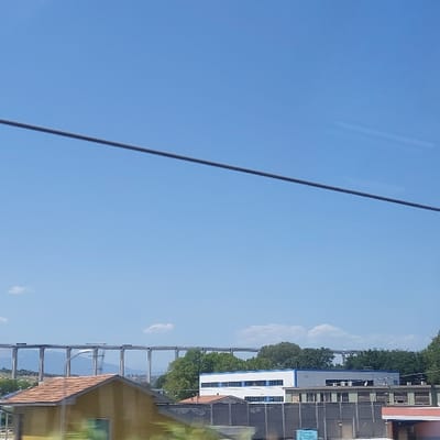 The image captures an outdoor daytime scene in the vicinity of Tortoreto, Italy, under a clear blue sky with sparse, high-altitude cloud formations. A thin, dark, horizontal cable or wire extends across the upper third of the frame. In the background, a large concrete viaduct, identified as a segment of the A14 Autostrada, stretches horizontally across the landscape, supported by numerous tall, cylindrical concrete pillars. Distant hills are visible beneath the left portion of the viaduct.

In the mid-ground, below the viaduct, a cluster of structures is present. A prominent, multi-story building with a white facade and a distinct row of blue-framed windows on its upper level is situated centrally. To its right, an adjacent grey-colored building is partially visible. A line of green deciduous trees is positioned behind these structures. A uniform grey barrier or wall runs horizontally across the lower mid-ground, obscuring the base of the buildings. In the lower left foreground, a portion of a building featuring a reddish-brown tiled roof and light-colored walls is visible. To the lower right, another building with a red-orange roof is partially discernible behind the grey barrier. No individuals or discernible actions are observed within the frame.