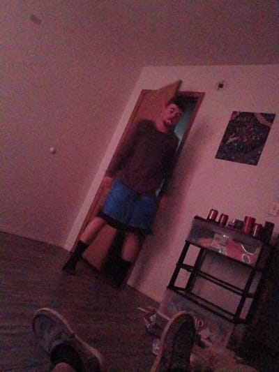 The image shows a room with a man lying in a doorway. He is wearing a dark shirt and blue shorts, with black socks. There is a picture on the wall above him, and a small black shelf with cans on it to his right. Two pairs of shoes are visible on the floor. The location is Clarksville, United States of America.
