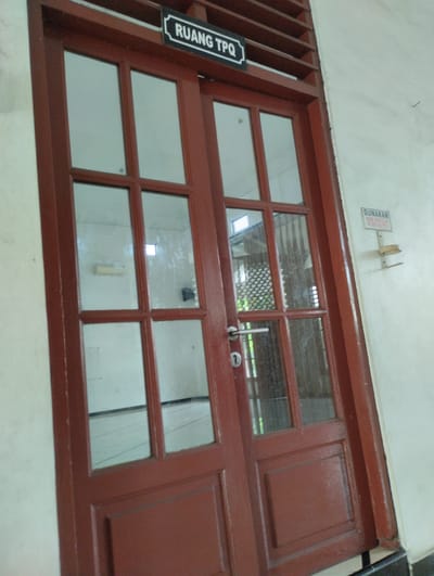 The image shows a reddish-brown double door with multiple glass panes and wooden panels. A sign above the door reads "RUANG TPQ" in white text on a black background. To the right of the door, on a white wall, is a sign that says "GUNAKAN HAND SANITIZER" with a hand sanitizer dispenser mounted below it. The door appears to be located in a building, possibly an educational institution, given the sign indicating "TPQ" (Taman Pendidikan Al-Qur'an), which is an Islamic learning center for children. The location is stated to be Tanjungsari, Indonesia. The environment appears to be indoors, with the reflection of lights and windows visible in the glass panes.