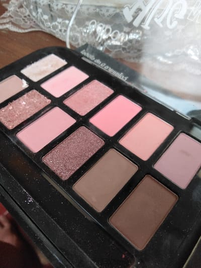 A close-up, high-angle forensic image, captured in Ciudad de San José, Costa Rica, depicts an open rectangular eyeshadow palette with a black plastic casing. The palette contains twelve individual eyeshadow pans arranged in three rows of four. The shades consist of various matte, shimmer, and glitter formulations in a color range dominated by pinks, browns, and nudes. Specific visible colors include: a disturbed light beige or off-white matte, multiple matte pinks, a brighter pink shimmer, a light peach matte, a light mauve matte, and several medium to dark brown matte shades. Two distinct glitter shades are present, one appearing rose gold and another a darker brown or bronze. Evidence of prior use is observed through powder fallout scattered across the black plastic casing, particularly along the left edge and between the pans. The top-left light beige pan shows significant surface disturbance, indicating frequent application. The background is blurred, revealing a white lace-like textile towards the upper right and a translucent plastic cover with illegible white script positioned above the palette. The surface directly beneath the palette appears to be a light brown, potentially wood.