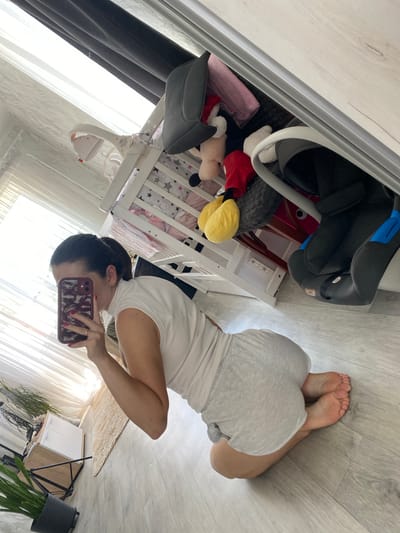 In Romny, Ukraine, a woman kneels on a light-colored wooden floor, facing away from the camera. She is taking a mirror selfie with her phone, which is held up to obscure her face. She is wearing a white sleeveless top and light grey shorts. Her hair is pulled back in a ponytail. To her right, a white crib with stuffed animals is visible. The crib appears to be filled with various plush toys, including characters resembling Mickey Mouse. A car seat is also present, positioned partially within the crib. In the background, sheer white curtains and darker drapes cover a window, and a dresser with drawers stands to the left. A plant in a dark pot is visible in the lower left corner. The lighting suggests it is daytime, with natural light streaming through the window.