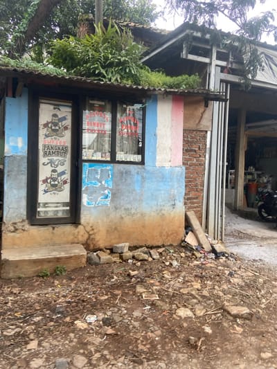The image shows a small storefront in Baleendah, Indonesia, located at latitude -7.022 and longitude 107.671. The storefront is a single room with a black door and two windows, both displaying signs advertising "Pangkas Rambut" (haircut) services. The room's exterior is painted blue and white with a pink stripe running vertically. The storefront is situated on a dirt path, with a small, concrete step leading up to the door. The ground in front of the storefront is covered in debris and rocks. A small pile of wooden planks is leaning against the side of the building, partially obscuring the entrance to a larger, open-air area. A motorcycle is visible in the background, partially obscured by a metal gate.  A small tree with green foliage overhangs the building from the left.