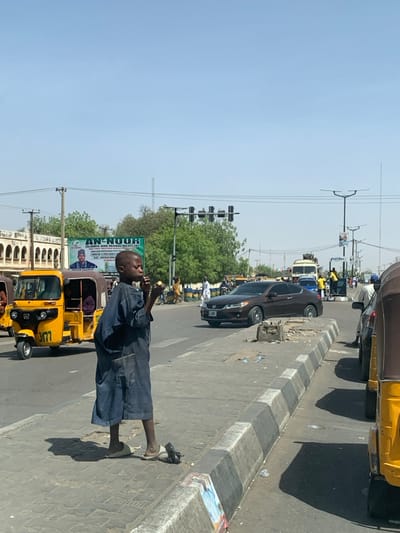 A young male individual, appearing to be a child, stands on a paved sidewalk or median strip on a sunny day in Hotoro South, Nigeria. He is wearing a dark blue, long-sleeved garment that extends below his knees and light-colored sandals. His right foot is in a sandal, while his left foot is positioned near a detached sandal and a dark object on the ground. He holds a spherical, light-colored object, possibly a piece of fruit, to his mouth with both hands, suggesting he is eating it. Strong shadows are cast to his left.
To the immediate left of the individual, a yellow three-wheeled auto-rickshaw with the number "3177" visible on its front is parked or moving slowly. Another similar vehicle is partially visible behind it. Across a paved street, a dark brown two-door Honda Accord coupe is in motion. Further down the street, additional vehicles including a truck and several people are visible.
In the background, a large green billboard displays "AN-NOOR ISLAMIC AND HERBAL MEDICINE" with an image of a man. To the far left, a light-colored building with multiple arched window or door frames is present. The sky is clear blue. Traffic lights are visible suspended above the intersection, alongside power lines and streetlights. A concrete median with alternating black and white stripes separates the central individual from the vehicles parked on the right side of the frame, which include the partial view of another yellow auto-rickshaw. Debris is scattered on the ground near the median.