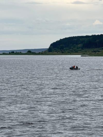 An eye-level, medium shot captures a wide, rippling body of water under a cloudy sky. In the middle distance, a small, dark inflatable boat with two occupants is moving across the water, leaving a slight wake. One person in the boat wears an orange life vest. The background features a tree-lined hillside with a darker band of forest along its top, and a strip of lighter green vegetation at its base, bordering the water. A hazy, light-colored band of land is visible further in the distance. The scene appears to be a natural landscape, likely a lake or a wide river, with no visible urban elements or specific landmarks indicating the city of Красный Бор, Russia.