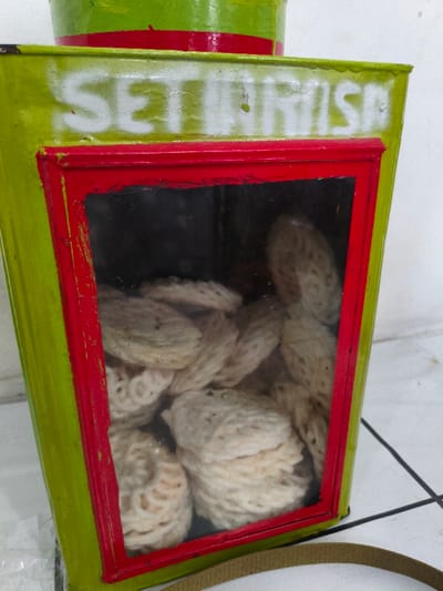 The image displays a brightly colored, rectangular snack container, located in Muka, Indonesia. The container is painted lime green, featuring a prominent red frame around a clear window on its front. Above this window, the white stenciled text "SETIA MISA" is visible. The upper part of the container is a cylindrical section, also green with a red horizontal band. Inside the clear window, numerous light-beige, textured, circular food items, consistent with krupuk or crackers, are tightly packed. The container is positioned on a white tiled floor with dark grout lines. A brown fabric strap is partially visible in the lower right foreground. The background behind the container is a plain, light-colored surface. No people or active interactions are visible in the scene.