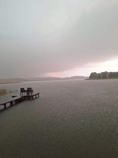 The image depicts a weathered wooden pier extending into a calm, dark body of water during a heavy rainfall. Two wooden chairs and a small table are situated on the pier, accompanied by a shovel leaning against them. The water's surface is visibly disturbed by numerous raindrops, creating a textured pattern. In the background, a distant, tree-lined shore is shrouded in mist and rain, blending into a grey, overcast sky. The overall atmosphere is somber and tranquil. The location is identified as Слабодкаўскі сельскі Савет, Belarus.