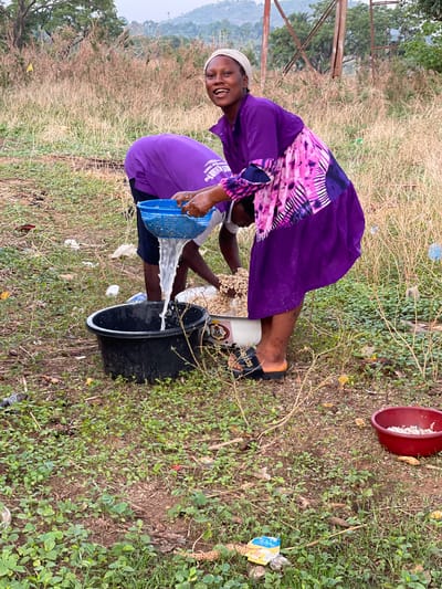 In an outdoor setting in Tabu, Nigeria, a woman is bent over pouring water from a blue plastic colander into a black plastic bucket. The woman is wearing a purple dress with a pink and white patterned top, a white head covering, and black Adidas slides. She is smiling and looking at the camera. Next to her is a white container filled with what appear to be nuts or seeds. A second person is partially visible behind the woman. A red bowl containing white matter is in the upper right corner. The ground is covered with a mix of brown grass, green foliage, and scattered trash. A hill with trees and power lines is visible in the background.