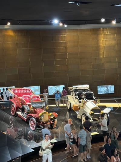This image depicts the Mercedes-Benz Museum in Stuttgart, Germany. The museum features a display of vintage automobiles, with two prominent cars in the foreground. To the left is a red open-top touring car with plush red seating and a spare tire mounted on the side. To the right is a white and cream open-top touring car with black seating and a small trunk mounted on the side. Several people are visible observing the cars. The background consists of a large, textured gold wall with several illuminated screens displaying historical imagery. The lighting is a mix of spotlights from above and ambient light from the screens. The floor is a dark, reflective surface with geometric patterns.