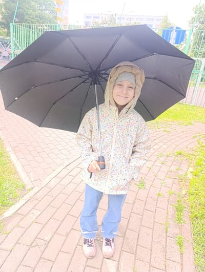 A young person stands on a brick walkway holding a black umbrella. They are wearing a floral jacket with a fur-lined hood, a gray hat, blue jeans, and pink sneakers. The background shows a grassy area, a metal fence, and apartment buildings, suggesting an urban environment. The location is Noginsk, Russia.