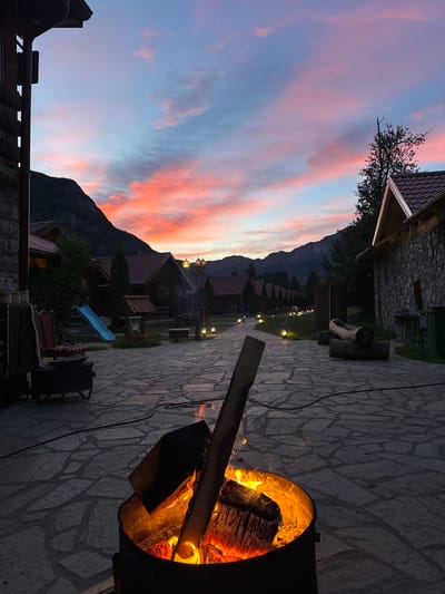 The image depicts an outdoor scene at dusk, likely in Gusinje, Montenegro, characterized by a vibrant sunset painting the sky in hues of orange, pink, and blue. In the foreground, a metal fire pit contains a glowing campfire with burning logs, casting a warm light on the surrounding flagstone patio. To the left, a portion of a building with a dark roof and stone base is visible. Further back, several wooden cabins with red roofs are arranged in a row, illuminated by small outdoor lamps. A blue slide for children is situated near one of the cabins. The background features a mountainous landscape under the colorful twilight sky. The overall impression is one of a tranquil evening in a mountain village setting.