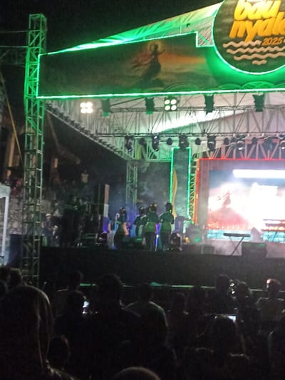 The image captures an outdoor night event, likely a concert or cultural performance, in Kecamatan Pujut, Indonesia. A large, complex stage structure, constructed from metal trussing, dominates the scene, bathed in bright green illumination.
On the stage, several performers are visible, some dressed in green attire, amidst stage lights and a light haze or smoke effect. The performers appear engaged in an act, suggesting an ongoing show. Above the main stage, a prominent rectangular LED screen displays a striking graphic: a silhouetted figure, possibly a dancer or mythical creature, against a vibrant, fiery background. To the upper right, a large circular banner or sign features the text "bau nyale 2025" with wavy lines underneath, indicative of a festival or event name scheduled for the year 2025. Another bright LED screen is positioned on the right side of the stage, also displaying visuals.
The foreground is filled with the dark silhouettes of a large audience, gathered to watch the performance. Their heads and shoulders are visible, indicating a significant crowd. Some audience members appear to be holding up phones, potentially recording the event or taking photos. The dark sky above confirms the nighttime setting, with all illumination originating from the stage lighting and screens. The overall impression is that of a lively public gathering for a cultural or entertainment event.