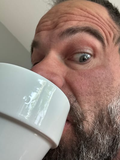 A close-up, tilted view shows a person's face, likely male, with graying hair and a beard. Wrinkles are visible on the forehead. The person's eye is visible, reflecting light. A large, white, cylindrical object, possibly a mug or cup, is positioned close to the person's face, partially obscuring the mouth. The background is a plain, light-colored wall. There is no information available to establish a connection between the image and the city of Marche-en-Famenne, Belgium.