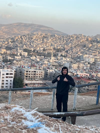 A panoramic image depicts a young male subject standing on an elevated overlook, smiling and presenting a "thumbs-up" gesture with both hands towards the viewer. He is dressed in a black hooded tracksuit and black sneakers with white soles. Immediately in front of him is a metal railing, consisting of an upper blue-painted bar and a lower black-painted bar, supported by vertical posts. The ground in the foreground consists of loose earth mixed with white and blue fibrous or plastic debris.

The midground showcases the densely populated urban landscape of Qudsaiya, Syria, extending across hilly terrain. Numerous multi-story residential and commercial buildings, predominantly light-colored, are tightly clustered. A prominent white mosque minaret is visible within the city.

In the background, a range of arid, rocky mountains stretches across the horizon. A cluster of communication towers is discernible on one of the distant mountain peaks. The sky is clear and light blue, indicating daylight conditions, likely late afternoon due to the soft, directional illumination on the city.
