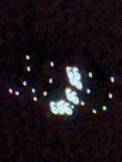 This is a grainy, low-light photograph taken in Stallings, United States of America, likely at night. The image depicts a cluster of bright, blurred white lights against a dark, textured background. Several of these lights are formed into the shapes of numbers. In the upper right portion of the image, the number "65" is visible, with the "6" appearing above and slightly to the left of the "5". Below and to the left of "65", another cluster of lights forms the number "666", with the three "6"s arranged in a somewhat triangular pattern, though the shapes are indistinct due to the image quality. Scattered throughout the scene are smaller, individual white lights, some appearing as pinpricks and others as slightly larger blurs. There are no visible people, animals, or discernible structures in the photograph. The overall impression is of a display of illuminated numbers or symbols against a dark, possibly outdoor, environment.