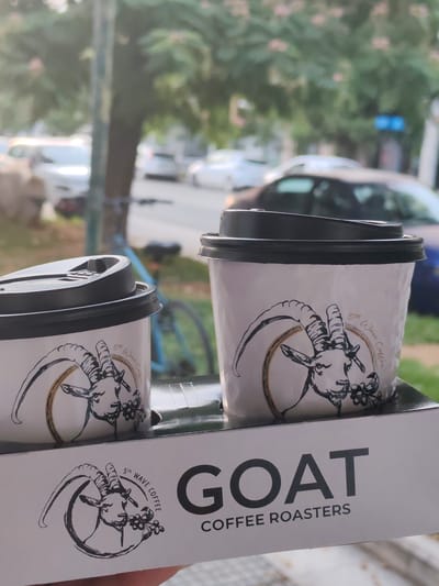 Two disposable coffee cups with black lids are placed in a white cardboard carrier. Both cups feature a circular logo with a black and white illustration of a goat's head and the text "5th Wave Coffee" around it. The carrier itself has the word "GOAT" printed in large black letters, with "COFFEE ROASTERS" written below it. The background shows a blurred street scene with cars and trees, suggesting an outdoor setting. The presence of the coffee cups and carrier indicates a purchase from a coffee shop. The reflection of what appears to be a leafy tree and sky on the surface of the cups and the nature of the objects suggest an outdoor seating area or a location near a window where light is reflecting. The overall image is slightly out of focus, with the coffee cups being the primary subject. The scene suggests a casual moment, likely a person holding the coffee carrier, which is partially visible in the foreground. The blurred background hints at an urban environment, possibly a street in Alexandroupolis, Greece.