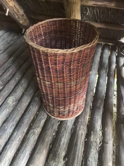 In the interior of a rustic structure, a wicker basket sits on a floor made of parallel wooden logs. The basket is woven from thin, flexible strands of a reddish-brown material, with a lighter colored rim. The logs are rough and weathered, with visible grains and occasional darker spots. Above the basket, the ceiling is constructed from similar wooden beams and a thatched material. The overall impression is one of natural materials and a simple, handmade construction, possibly in a rural or historical setting. The location of Камянюцкі сельскі Савет, Belarus, suggests this could be a traditional dwelling or outbuilding in a Belarusian village. No people are present in the image.