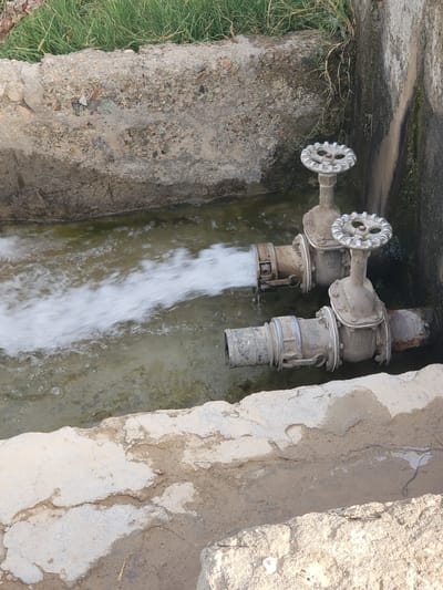 The image shows two pipes expelling water into a concrete channel. The pipes are equipped with large, round valves. The surrounding environment consists of concrete structures and some vegetation. The location is possibly in Arghandab, Afghanistan, as requested, but this is not ascertainable from the image alone. No people are visible.