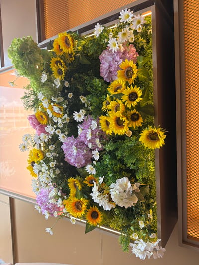 A low-angle shot of an indoor floral display is framed by a rectangular structure, possibly mounted on the ceiling. The arrangement includes sunflowers, white daisies, purple and pink hydrangeas, and green foliage. The structure has a dark brown or black frame with a mesh-like material above the flowers and a reflective surface below, possibly glass or polished material. The environment suggests a commercial or public space. The city of Torreforta, Spain, cannot be determined from the image.
