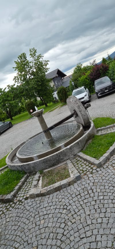 The image depicts an outdoor scene in Eugendorf, Austria, featuring a stone fountain as the central element. The fountain is circular with a tiered design, and water is actively flowing from a central spout into the basin. A large, weathered millstone is positioned at the edge of the fountain, partially submerged in water, and connected to a central pillar by a wooden beam. The ground surrounding the fountain is paved with cobblestones, and there are grassy areas and some planted bushes and trees. In the background, several vehicles are parked on a paved surface, and a building with a dark roof is visible. The sky is overcast with dramatic clouds. A dark gray sedan is parked to the left of the fountain, and two other cars, a white SUV and a dark SUV, are parked in the background. The overall impression is of a tranquil, slightly rustic setting in a residential or public area.