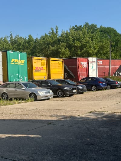 In Schkopau, Germany, a row of parked cars is visible in front of several shipping containers. The cars, including a silver station wagon, a black sedan, a dark sedan, and a blue SUV, are lined up on a paved surface. Behind the cars, large shipping containers are stacked. Some containers are yellow and display logos such as DHL and "FRYE INT. SPEDITION". Others are red and white, with text including "TRANS COR" and "KRISSEL TRANSPORT". The background consists of a line of green trees against a clear blue sky. The ground in the foreground is a mix of concrete and asphalt, with some patches of grass. The scene appears to be an outdoor storage or logistics area.