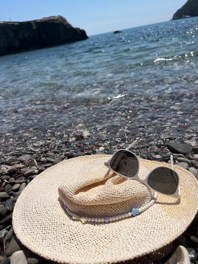 In Zagrađe, Montenegro, a straw hat and sunglasses lie on a pebble beach. The beach is adjacent to the clear, blue water of the Adriatic Sea, which reflects the bright sunlight. In the background, a dark, rocky outcrop juts out from the sea. The hat is light beige and has a decorative string of white and pastel beads with a blue flower charm. The sunglasses are white with dark lenses. The scene depicts a serene beach setting on a sunny day.