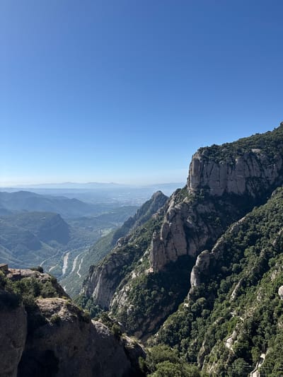 The image depicts a dramatic, high-angle view of a mountain landscape, likely taken from a significant elevation. The foreground is dominated by a sheer, rocky cliff face covered in dark green vegetation. This cliff drops away sharply, revealing layers of receding mountain ranges covered in dense foliage, fading into a hazy blue sky. A winding road is visible in the valley below, snaking through the forested slopes. The sun appears to be high in the sky, casting strong shadows and illuminating the scene with bright light, suggesting it is daytime. The clear blue sky indicates fair weather. The scale of the mountains and the vastness of the vista evoke a sense of awe and perhaps a touch of vertigo. While no people are visible, the presence of a road and what appear to be structures or trails higher up on the mountainsides suggest human presence or activity in the area. The location is identified as Monistrol de Montserrat, Spain, a region known for its distinctive rock formations and mountainous terrain. The overall impression is one of natural grandeur and a remote, rugged environment.