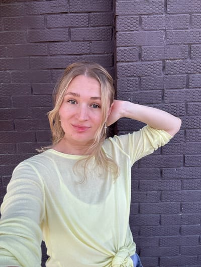 A young Caucasian woman with blonde hair is shown from the chest up, posing for a selfie in front of a dark purple brick wall. She is wearing a light yellow long-sleeved shirt that is tied at the bottom. Her right arm is bent at the elbow and her hand is resting on the back of her head. She is looking directly at the camera with a slight smile. The brick wall has a hexagonal pattern. The image does not depict the city of Verkh-Sosnova, Russia.