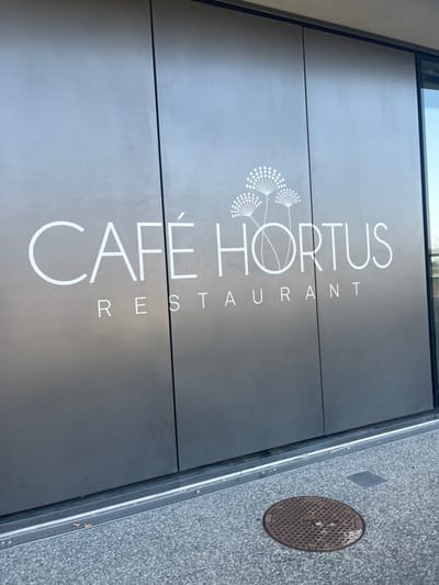 The image captures the modern facade of a restaurant named "Café Hortus" in Meyrin, Switzerland. The building entrance features a dark, possibly grey or black, surface adorned with prominent white lettering and a distinctive graphic. The main name "CAFÉ HORTUS" is displayed in large, sans-serif capital letters, with an acute accent visible over the 'E' in 'CAFÉ'. To the left of the 'H' in 'HORTUS' is a stylized white graphic depicting three delicate, circular forms reminiscent of dandelion seed heads. Below the main name, the word "RESTAURANT" is spelled out in smaller, spaced-out capital letters.

To the right of the building, a grey paved area, likely a sidewalk, is visible. It features a circular metal manhole cover with a hexagonal grid pattern. The scene is illuminated by natural light, indicating daytime, with the uniform lighting suggesting an overcast sky or indirect sunlight. There are no people or activities taking place, presenting a static view of the restaurant's exterior.