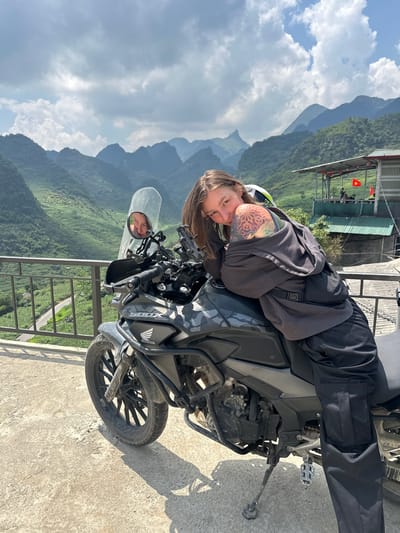 A woman with brown hair and a tattoo on her left arm is reclining on a gray and black Honda CB500X motorcycle. The motorcycle is parked on a concrete platform near a black metal railing overlooking a mountainous landscape. The location is Lũng Phìn Commune, Vietnam, as indicated by the scenery. Behind the motorcycle and railing, there are green mountains and a cloudy sky. The woman is wearing a gray hooded sweatshirt and black pants. In the motorcycle's windshield, a reflection of the woman's face can be seen. In the background, there is a structure with a green roof and a Vietnamese flag.