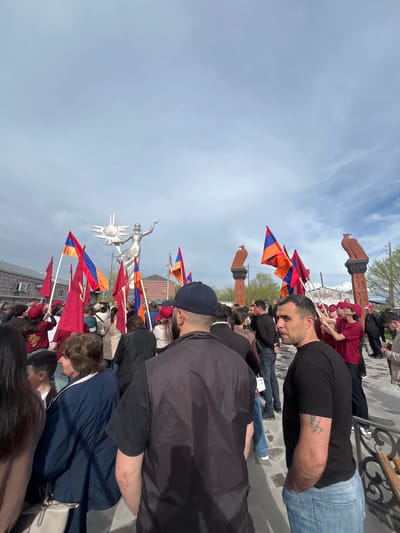 A crowd of people hold Armenian flags and congregate in a public square in Akhuryan, Armenia, near the Mother Armenia statue. The sky is partly cloudy. In the foreground, there are many people wearing casual clothing, some wearing red shirts. Flags are waved by some of the people in the crowd. The Mother Armenia statue is in the middle of the shot and is light-colored. There are also decorative columns with avian figures.