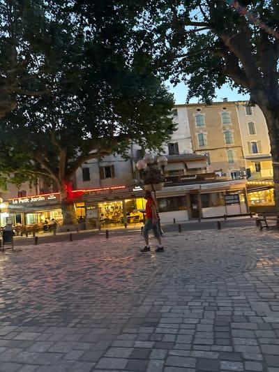 This image depicts a street scene in Anduze, France, likely taken during dusk or evening, as indicated by the artificial lighting and the twilight sky. In the foreground, a person with long, light brown hair, wearing a red t-shirt and gray shorts, walks across a cobblestone plaza. They are moving from left to right, with their body slightly turned towards the viewer.

Behind the pedestrian, a row of illuminated restaurants and cafes line the street. A prominent sign reads "Le Petit Jardin," above which is a red neon light. Other establishments are visible, with tables and chairs set up outside. The buildings are typical of European architecture, with multiple stories and visible windows. Large trees with dense foliage are scattered throughout the scene, providing shade and framing the buildings. The cobblestone paving in the plaza has a distinct circular pattern in some areas. The overall atmosphere suggests a lively evening in a town square, with people dining and socializing outdoors.