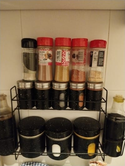 A three-tier black wire spice rack hangs on a white tiled wall. The bottom tier contains three black jars with white speckled lids and clear rectangular windows exposing their contents. Bottles of dark liquid in similar holders flank the spice jars. The middle tier mirrors the bottom with five speckled-lidded jars. On the top tier, a row of five spice bottles in plastic containers sit. From left to right, the lids are black and red. The labels and contents of the plastic bottles are partially visible.