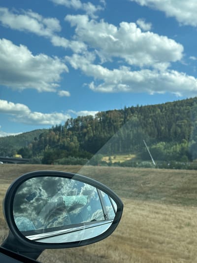 The image shows a side-view mirror of a car, reflecting a cloudy blue sky and a wooded hillside. The car is on a road with dry, golden grass on either side. In the distance, a highway overpass can be seen. The image appears to have been taken on a sunny day. There is no visible indication of the city of Haslach im Kinzigtal, Germany, within the frame of the image.
