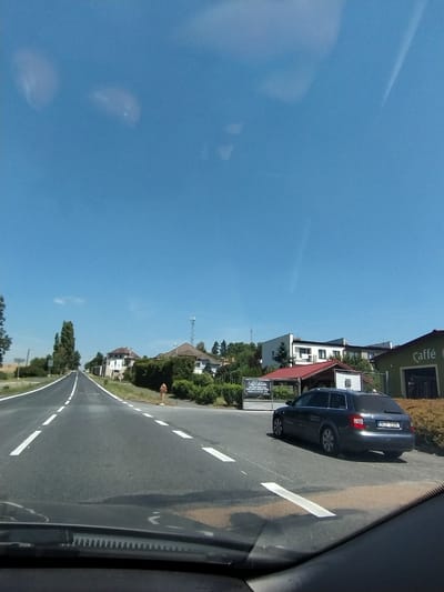 The image depicts a sunny day in Horažďovice, Czechia. The scene is viewed from a car, looking out at a road winding through a village. The sky is a clear, vibrant blue, suggesting pleasant weather.
On the right side of the road, several buildings are visible, including what appears to be a cafe with the word "Caffè" prominently displayed. A dark-colored car is parked by the side of the road. Signs with text, including what seems to be an advertisement for "Caffè Edelsitz," are present. The architecture of the buildings suggests a European setting.
The road itself has white dashed lines marking the lanes, indicating it's a paved road. The surrounding area is green, with trees and grass lining the sides of the road, further contributing to the impression of a rural or suburban village.
In the distance, a telecommunications tower can be seen, adding to the sense of being in a populated area. The overall impression is of a calm, ordinary day in a picturesque Czech village. The time of day appears to be midday, given the brightness of the sun and the clear blue sky. No people are clearly visible in the frame, but the presence of parked cars and buildings suggests ongoing activity.