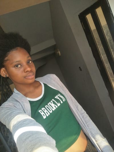 A young individual with dark skin and dark, textured hair styled upwards is captured in a head-and-torso selfie. The subject wears a dark green sleeveless top with white trim and "BROOKLYN" printed in white capital letters across the chest, partially covered by a light grey open cardigan featuring two white horizontal stripes on the visible left sleeve. A small, dark stud earring with a central metallic detail is visible in the left ear. The subject's expression is neutral, with eyes directed towards the camera and lips slightly parted. The background consists of uniformly dark grey interior walls. To the upper right, a dark-framed door or window with multiple vertical glass panels is visible. A circular, light-toned object, consistent with a wall-mounted light fixture, is present on the wall to the upper left. Subtle dark, vertical railing elements are discernible in the lower-left background. The scene is illuminated by ambient light, with a brighter illumination source from the right side of the frame. The specific location within Cotonou, Benin, cannot be determined from the visual evidence presented.