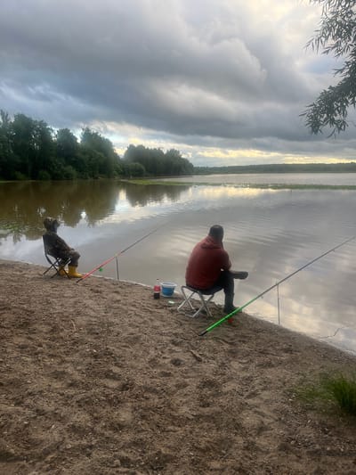 The image depicts two individuals fishing by a calm body of water. The scene is set on a sandy shore, with lush green trees lining the opposite bank of the water. The sky is overcast with dramatic clouds, suggesting it might be late afternoon or early evening, with a hint of sunlight breaking through on the horizon.
One person, wearing an orange hoodie and dark pants, is seated on a folding chair, holding a fishing rod. Another person, camouflaged in what appears to be fishing attire and yellow boots, is also seated on a folding chair and holding a fishing rod. Their fishing rods are propped up, indicating they are waiting for a bite. Various fishing gear, including a bucket and a bottle of Coca-Cola, are placed near the shore.
The overall atmosphere is one of peaceful recreation. The calm water reflects the sky and trees, creating a serene and natural environment. The location is identified as Klepiki, Russia. There is no visible text in the image.