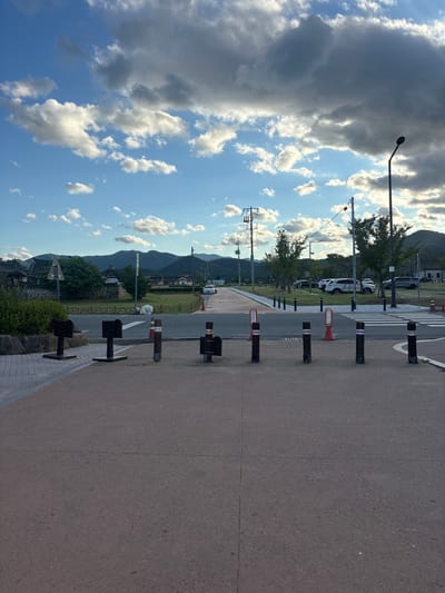 The image depicts a scene in Gyeongju-si, South Korea, featuring a road intersection with a pedestrian crossing. In the foreground, there are dark bollards placed on a paved walkway. The road itself is asphalt with white lane markings and a pedestrian crossing.

Further into the scene, a white car is visible on the road. To the left of the road, traditional Korean-style buildings with dark tiled roofs are seen, surrounded by stone walls and green foliage. In the background, a line of mountains forms the horizon under a partly cloudy sky with dramatic cloud formations.

A streetlamp with a black fixture is present on the right side of the image. Several other dark bollards are positioned along the roadside and near the parking area. The environment suggests a historical or cultural site, given the architecture. The lighting indicates it is either early morning or late afternoon. There are no people visibly present in the scene.