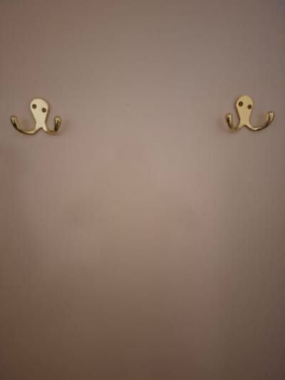 The image depicts a wall with two gold, octopus-shaped coat hooks. The wall is a light, neutral color, possibly beige or taupe. The hooks are mounted on the wall, one on the left and one on the right, positioned at roughly the same height. Each hook has a round head with two small, circular indentations resembling eyes, and two curved arms extending downwards, designed to hold coats or other items. The lighting in the image is soft, casting a subtle shadow around the hooks. The background is smooth and unadorned. There is no information provided in the image or accompanying text to indicate the location of this scene within Huntingdonshire, United Kingdom.