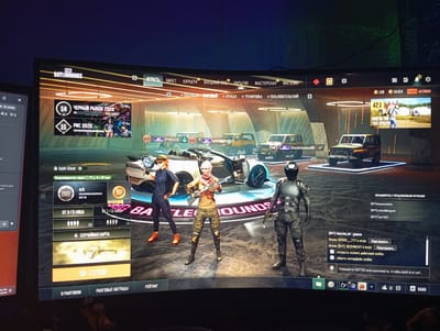 The image displays a computer screen showing a video game interface, specifically a lobby screen for "PUBG BATTLEGROUNDS". The game is presented in a first-person perspective. On the left side of the screen, there's a window from another application displaying game-related information, such as chat messages and player counts. The main game screen features three characters in combat gear, two females and one male, standing in a futuristic, well-lit environment with a large car in the background. The interface includes various game menus and options, such as "PLAY", "TICKET", "CAREER", "SETTINGS", "WORKSHOP", and "STORE", with the "PLAY" button prominently displayed. There are also indicators for player levels, in-game currency, and various game modes. The overall scene is a digital representation of a video game environment. The image does not contain any elements related to Sanremo, Italy.