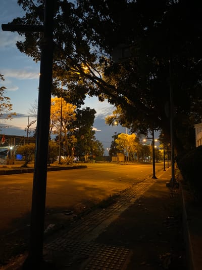 The image displays an urban street intersection in Parungserab, Indonesia, observed at dusk or early night. The scene is illuminated primarily by warm, yellow streetlights, creating a strong contrast with the fading blue and grey sky overhead. The asphalt road is clear of vehicles and pedestrians, with light reflections indicating its surface. A dark, vertical pole, likely a streetlight post, is prominent in the immediate left foreground. A paved sidewalk, featuring a section of tactile paving, runs along the right side of the road. Dense trees and foliage line both sides of the street, with some leaves glowing yellow from the artificial light while others remain in silhouette. Multiple streetlights on black poles are visible extending into the distance along the roadside. On the far left, beyond the road, a structure with a red flag and a blue tarp is partially discernible amidst more trees. No human figures or active elements are present.