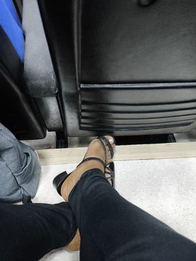 The image, taken in Kızılbaş, Northern Cyprus / Cyprus, shows a low-angle view from a seated perspective. In the foreground, a person's legs, clad in black trousers, are crossed at the ankles. The visible foot wears a black open-toed sandal featuring multiple thin, textured straps across the forefoot and around the big toe, with a low block heel. The skin on the exposed foot and ankle is tan. To the immediate left, a portion of a grey fabric backpack is visible on the floor. A light-colored wooden or plastic plank runs horizontally across the floor in front of the person's feet. The floor is light-colored and speckled. In the background, the lower rear sections of two dark, textured seats, likely synthetic material, are visible, featuring horizontal ribbed detailing. A dark vertical support structure is discernible between the two seats.