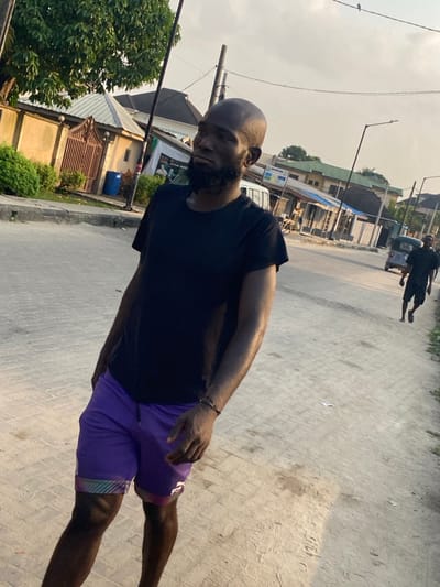 A bald, bearded man in a black t-shirt and purple shorts stands on a paved road. The photo is taken from a slightly high angle. A man in dark clothing runs in the background. Buildings and trees line the road. The sky appears hazy. The man in the foreground has dark skin and appears to be outdoors during the day.