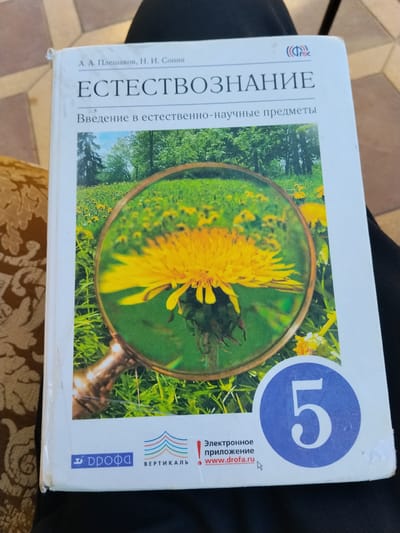 A close-up, overhead shot captures a book titled "ЕСТЕСТВОЗНАНИЕ" (Natural Science) displayed on a dark fabric surface. The book's cover features a vibrant image of a dandelion magnified by a magnifying glass against a backdrop of a sunny meadow. The title, authors' names (А. А. Плешаков, Н. И. Сонин), and a subtitle ("Введение в естественно-научные предметы") are visible in Russian. The book also shows publisher logos, a digital application notice with a website address (www.drofa.ru), and the numeral "5" within a blue circle. The overall scene suggests a controlled, indoor setting, possibly a desk or a lap, with the book being the sole focus. There is no discernible information or context within the image to associate it with the city of Dagestanskiye Ogni, Russia, or any specific urban environment.