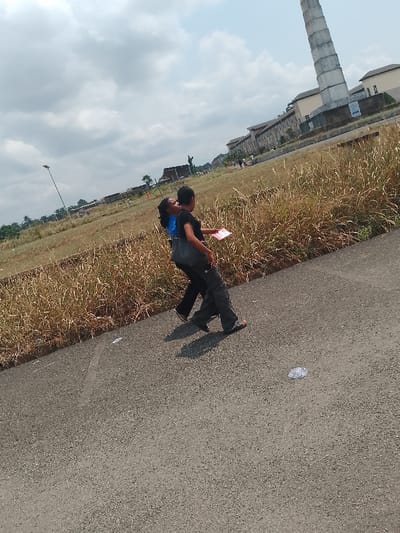 Two people walk together on an asphalt road with grassy areas on the side, near the National War Museum in Umueshi, Nigeria. The person on the left wears a blue shirt and dark pants, while the person on the right wears a black top, gray pants, and carries a dark-colored bag over the shoulder. The person on the right holds a pink object in hand. A tall chimney-like structure is visible in the background, as well as other buildings. Litter is scattered on the road.