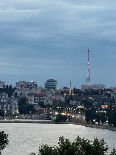 The image shows a cityscape at dusk, viewed from a high vantage point overlooking a river. The sky is a pale, overcast blue, suggesting either early morning or late evening. On the left side of the frame, a tall, lattice-like tower with red and blue lights stands prominently against the sky. The city itself is spread out along the riverbank, with numerous buildings of varying heights and architectural styles. Many of the buildings have illuminated windows, indicating that it is nighttime or twilight.
A road runs along the river, with cars visible, their headlights creating streaks of light. The river is wide and appears calm, reflecting the ambient light. Trees are visible in the foreground on the right side of the image, partially obscuring the view of the city and river. The overall impression is one of a sprawling urban environment transitioning into night, with the artificial lights of the city beginning to dominate the natural light. The scene conveys a sense of urban activity and the quiet transition from day to night.
There is no visible text in the image. The location context is Alexandrovka, Russia.