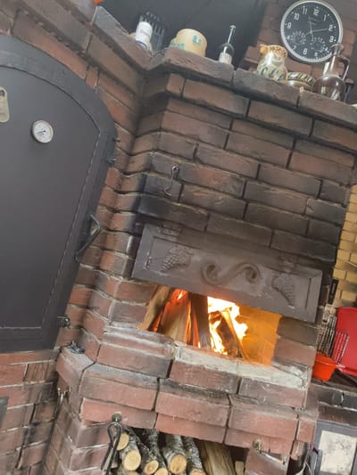 The image depicts a close-up, slightly angled view of a brick oven with a fire burning inside. The oven door, made of dark metal, is on the left side of the frame and features a circular thermometer. Above the oven opening, a decorative metal plate with grapevine motifs is visible. The bricks are reddish-brown and appear weathered, with some darker, soot-stained sections, particularly around the oven opening.
On the mantelpiece above the oven, several objects are displayed. These include a round, black clock with white numbers and hands, a ceramic pitcher with a yellow and blue pattern, a glass bottle with a dark label, and a brown ceramic jug. A small can with a white and blue label is also present.
To the right of the oven opening, a stack of chopped firewood is visible. Next to the firewood, a metal fire poker with a hook is propped against the brickwork. Further to the right, a red plastic bucket and a metal grill basket are partially visible.
The background is a brick wall. The lighting suggests an indoor environment, possibly a kitchen or a rustic dining area. The overall impression is one of a functional and decorative oven, likely used for cooking, situated in a room with a rustic aesthetic. The location is specified as "Востец Снт, Russia," which is an abbreviation for "Vostets SNT," likely referring to a horticultural non-profit partnership in Vostets, Russia.